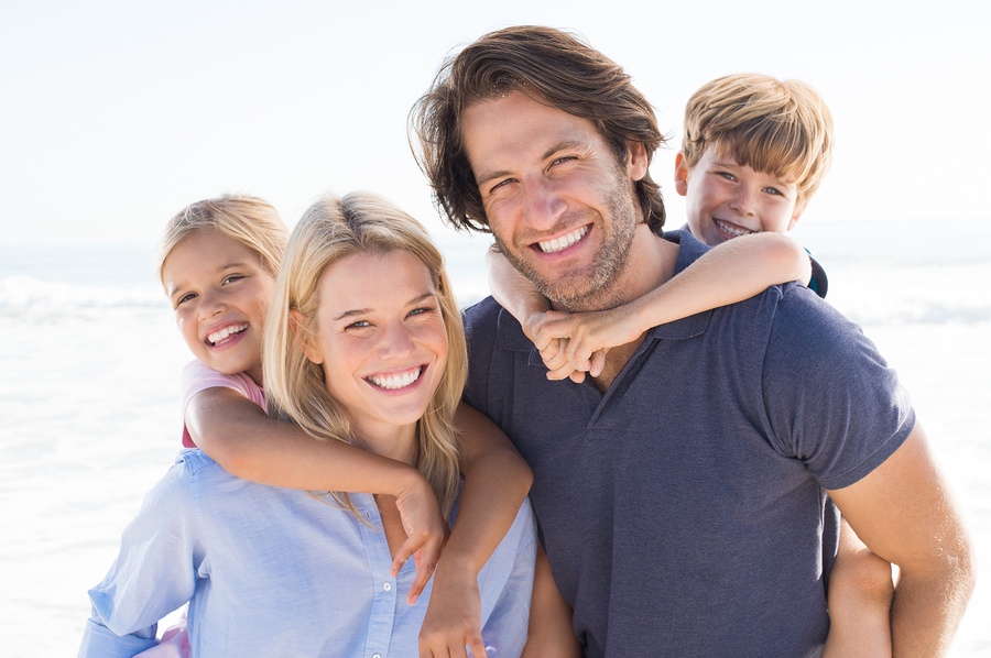 Parents giving piggyback ride to kids at beach. Close up of smiling family having fun at summer vacation. Portrait of happy family looking at camera at beach.